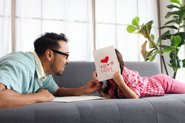 Happy Father Day, cute little girl with young man dad lying on sofa in living room, making and giving handmade greeting card for calibration. Lovely family daughter and father spending time together.