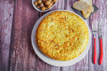 typical spanish potato omelette freshly made on a wooden table with cutlery, bread and olives.
