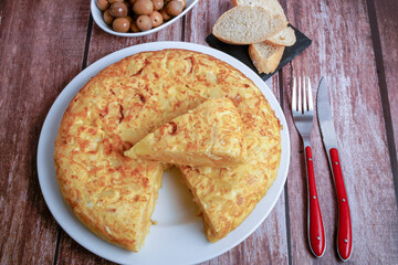 typical spanish potato omelette freshly made on a wooden table with cutlery, bread and olives.