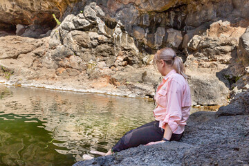 A young  girl sits on a stone on the shore and wets her feet in the Zavitan stream in the Yehudia National Natural Park in northern Israel