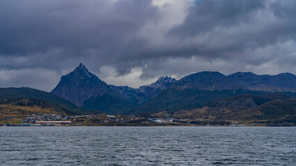 Picturesque snow-capped Martial mountain range against a cloudy sky. City houses are visible in the valley. In the foreground is the blue water of the Beagle Canal. Ushuaia. Argentina.