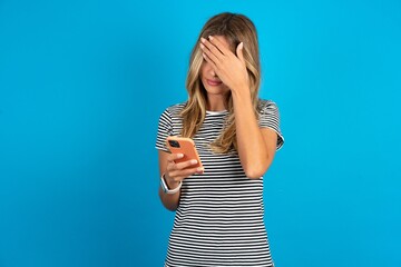 young beautiful woman wearing striped t-shirt looking at smart phone feeling sad holding hand on...