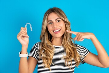 Young beautiful woman wearing striped t-shirt holding an invisible aligner and pointing perfect straight teeth. Dental healthcare and confidence concept.