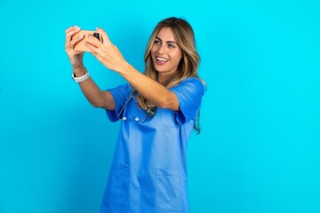 Young caucasian blonde doctor woman wearing blue uniform taking a selfie to post it on social media...