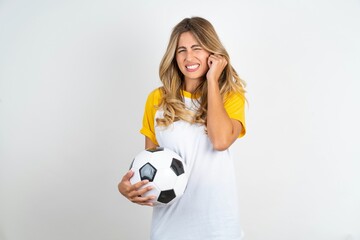 Happy Young beautiful woman wearing football T-shirt over white background ignores loud music and plugs ears with fingers asks to turn off sound