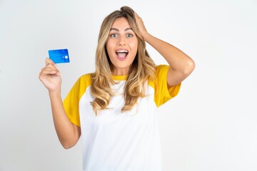 Impressed funny Young beautiful woman wearing football T-shirt over white background hand on head holding bank card