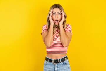 Young beautiful blonde woman wearing striped t-shirt over yellow studio background with scared...