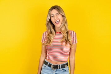 Young beautiful blonde woman wearing striped t-shirt over yellow studio background winking looking...