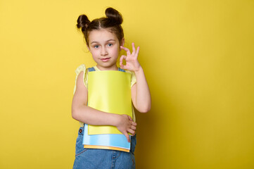 Cute lovely clever school girl, first grader shows OK hand sign with her fingers, smiles looking at camera, enjoying the start of a new semester of academic school year, isolated on yellow background