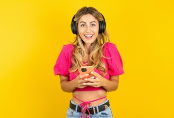 Excited young blonde woman wearing pink crop top over yellow studio background holding smartphone...