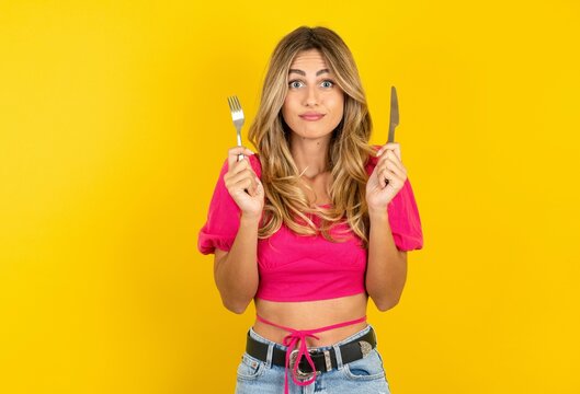 hungry young blonde woman wearing pink crop top over yellow studio background holding in hand fork knife want tasty yummy pizza pie