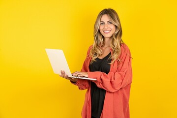 Adorable young beautiful blonde woman standing over yellow studio background holding modern device
