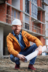 Cheerful man in protective work helmet holding architectural building plan and smiling while...