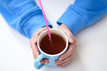 Kubek z gorącą herbatą trzymany w dłoniach, różowa plastikowa słomka jednorazowego użytku w...