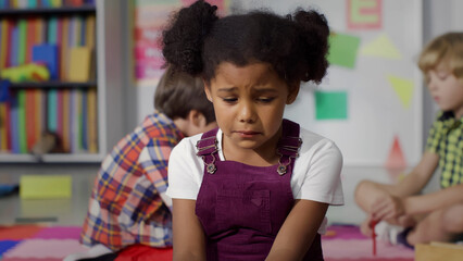 Close-up shot of upset African-American girl crying while playing with another kids in kindergarten