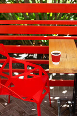 A wooden table on which there is a red plastic cup with coffee. Summer beautiful terrace in greenery. Cozy place.