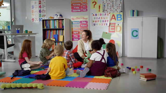 children sitting on floor while caring teacher explains lesson using toy in kindergarten
