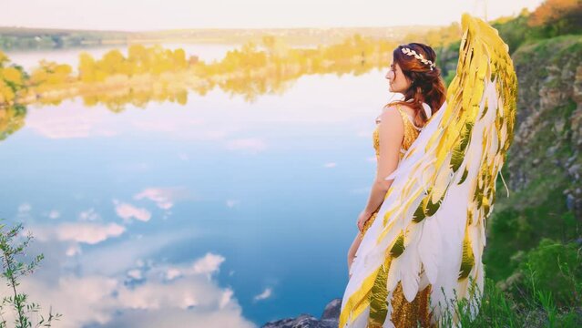 Happy fantasy woman angel with golden white wings walks on top of mountain magic blue sky green grass hills. Long dress girl greek goddess. Fairy queen sexy fairy lady. Summer nature water lake art 4k