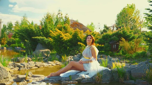 Fantasy woman goddess river nymph sits on stone rock lake. golden crown gold metal jewelry set Greek style. Queen girl fashion model in white dress enjoy summerr nature water divine sun magic light 4k