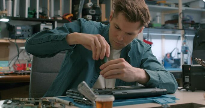 serious man disassembles laptop. Computer service and repair concept. Laptop disassembling in repair shop, close-up. Electronic development, device fixing. Master unscrews detail of broken computer