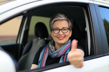 Cheerful senior woman in glasses sits in the driver's seat and shows thumbs up.