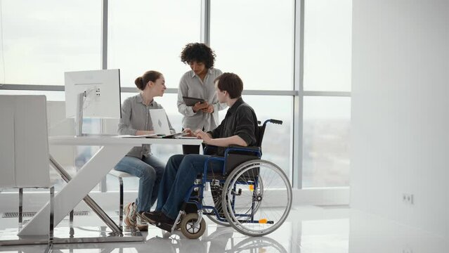 Young male office worker in a wheelchair talking to female colleague while showing her ideas using laptop in co-working space. Disability and handicap concept. Office modern people at work.
