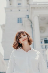 Wind hair style. A portrait of a woman outdoors, her shoulder-length brown hair blowing in the wind. Dressed in a white shirt against a light building.