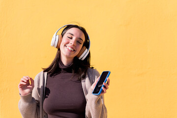 woman dancing happy listening to music from phone in headphones with a yellow wall in the...