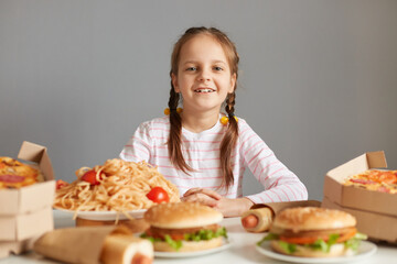 Smiling positive little girl with braids sitting at table with junk food isolated over gray background enjoying to eat unhealthy meals looking at camera with happy face.