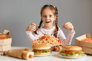 Amazed excited little girl with braids sitting at table with junk food isolated over gray background holding forks eating pasta looking at camera with big eyes.