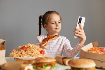 Cute little girl with braids sitting at table with junk food isolated over gray background holding slice pf pizza making selfie or video call on mobile phone.