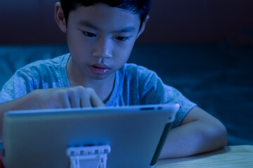 Background of Asian little boy age 7 years old watching a tablet and searching content on social media with dark low light in the bedroom and blue lighting reflection on his face. Selective focus.