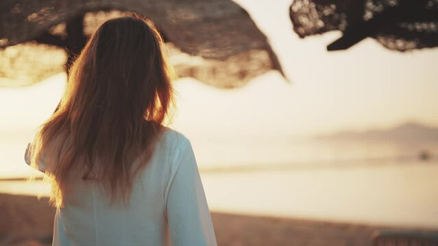 Girl resting walking to ocean on sandy shore under beach umbrellas on vacation at sunset touching hair, rear view. Woman relax near sea admiring amazing seascape at sundown. Travel, tourism concept.