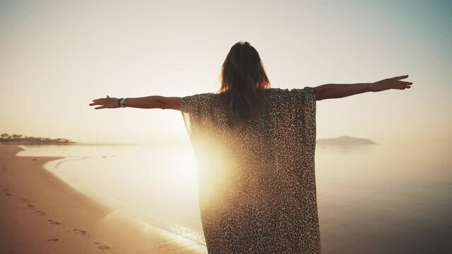 Woman in leopard pareo with arms stretched by sides stands at ocean sandy beach admires nature at sunset, back view. Freedom, carefree pose of relaxed girl near sea. Travel, tourism, enjoy vacation.