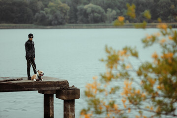 A man and an American Staffordshire Terrier stand on a pier by the river in autumn. A guy...