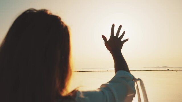 Woman touching sun in sunset sky over ocean enjoying vacation, rear view. Female feeling harmony, unity with nature looking at sunlight. Travel, tourism, summer holidays, resting outdoors concept.