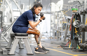 Woman with prosthetic leg sitting in gym lifting dumbbell weight. Asian female with foot prosthesis...