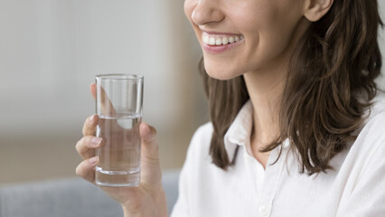 Close up cropped face view of beautiful happy young female holding glass of still or mineral water,...