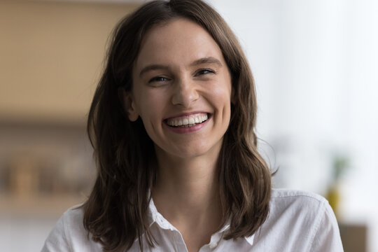 Close up head shot portrait of merry good-looking brown-haired young woman having wide-toothed candid smile staring at camera standing alone indoors, having attractive appearance and charming smile