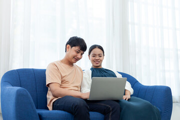 Close-up of Asian couple and pregnant woman using laptop to watch movies on blue sofa in living room.