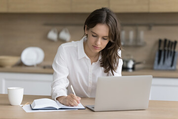 Attractive focused young woman takes notes, sitting at kitchen table with laptop, working or...