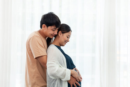 Close Up - Smiling Asian Man Wearing A Brown Shirt Hugs A Pregnant Woman In Blue. White Curtain Background.