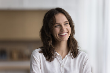 Head shot of beautiful calm woman standing alone in kitchen smile looking aside enjoy daydreaming on weekend at own or rented house. Millennial generation person portrait, happy mood and aspirations