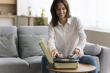 Happy woman placing vinyl record on turntable seated on couch, spend pleasant leisure resting at home, play nostalgic music, enjoy favourite melody, tune from the past. Retro lover pastime and hobby