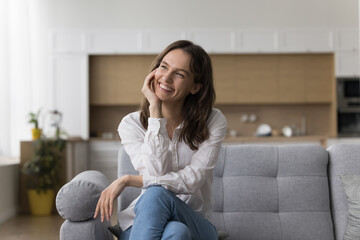 Cheerful woman finishes housework resting seated on sofa smile deep in pleasant thoughts, enjoy...