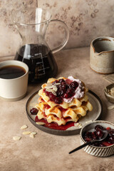 Belgian waffles with sour cream, cherry jam and almond petals topping and coffee on beige kitchen background. Sweet morning breakfast