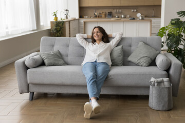 Millennial woman in casual clothes lying down on sofa with hands behind head smile feel happy on weekend at smart home with climate control enjoy cool air in summer day in studio apartment. Relaxation - obrazy, fototapety, plakaty