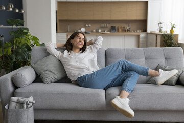 Cheerful millennial woman relax on sofa in studio apartment, leaned on soft comfortable couch with...
