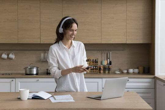 Woman In Headphones Holds Modern Phone Looks At Laptop Screen Spend Time In Kitchen At Home, Laugh, Listen Favourite Music Playlist, Enjoy Audio Book, Use Mobile Application For Study Foreign Language