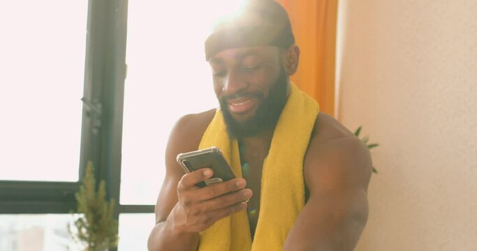sexy shirtless African American man with yellow towel on shoulder chatting with friends focused young male wearing sportswear, shorts using exercise bike typing message on phone. interests leisure